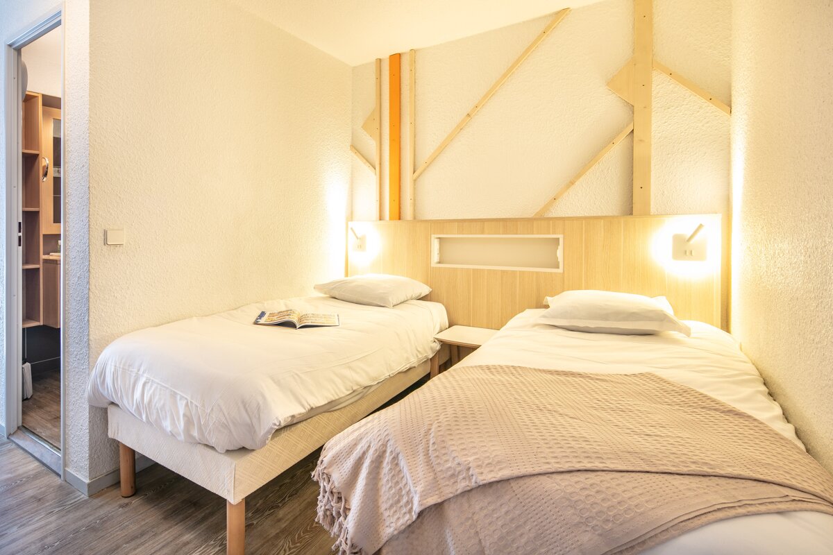 Bright bedroom with two single beds, light wood headboard with built-in lights, and textured walls with geometric wooden accents. Adjacent room seen.