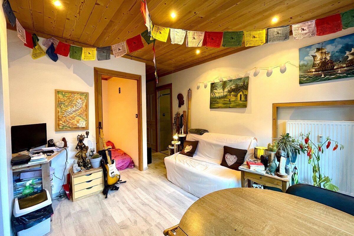 A cozy, eclectic room with a wooden ceiling, colorful prayer flags, a white sofa, various paintings, a guitar, and a desk area.