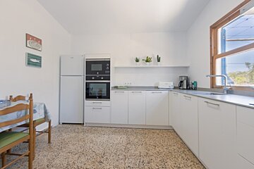 A bright, modern kitchen features white cabinets, built-in appliances, a corner sink by a window, and a small dining table on the left.
