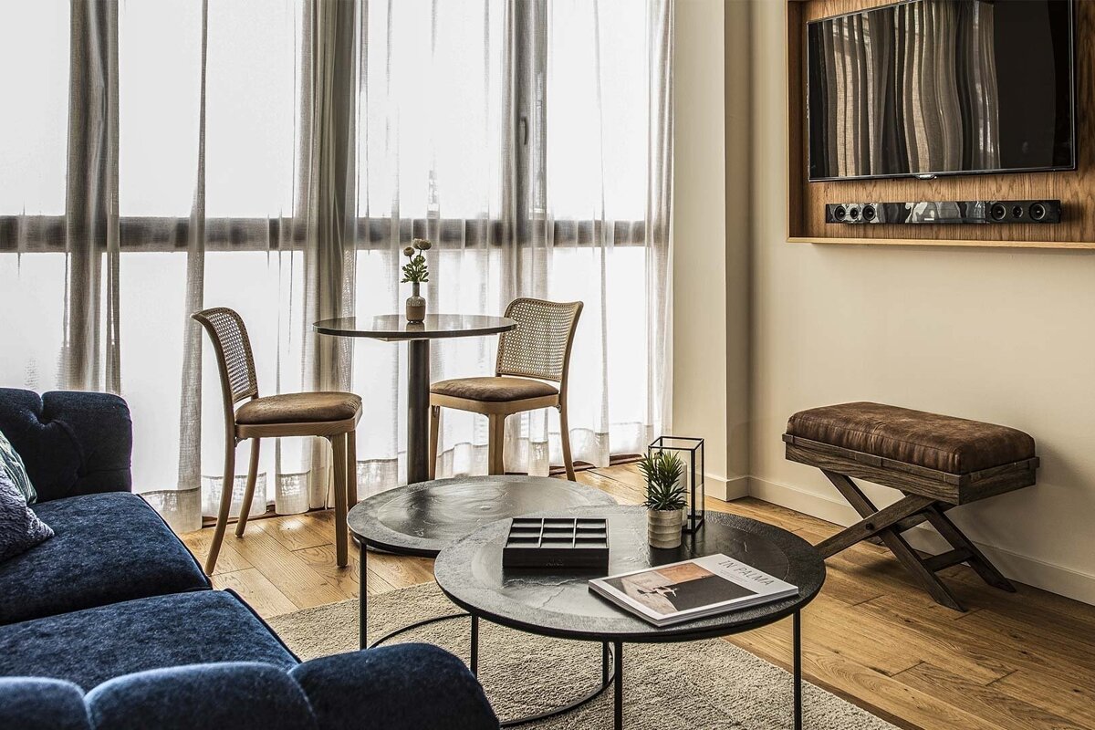 A stylish living space with a navy sofa, round coffee tables, a dining nook by a window with sheer curtains, a TV, and a leather bench.