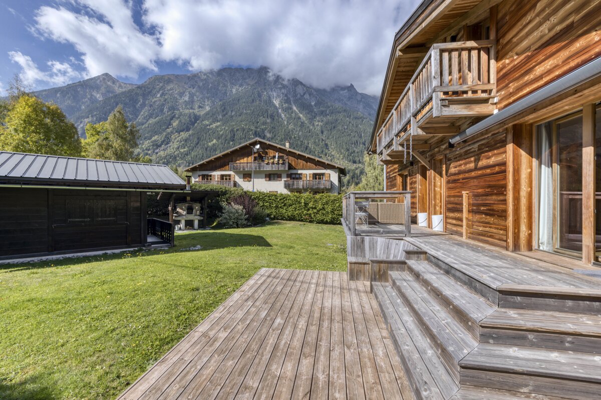 A rustic wooden chalet with a large deck and green lawn, set against a stunning backdrop of majestic, cloud-covered mountains and other houses.