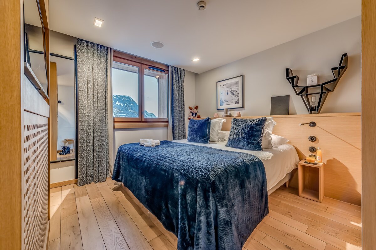 A stylish bedroom with a large blue bed, light wood flooring, and a window revealing a beautiful mountain landscape. Modern decor and cozy ambiance.