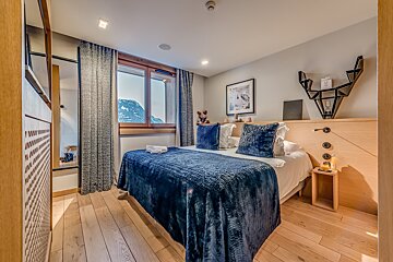 A stylish bedroom with a large blue bed, light wood flooring, and a window revealing a beautiful mountain landscape. Modern decor and cozy ambiance.