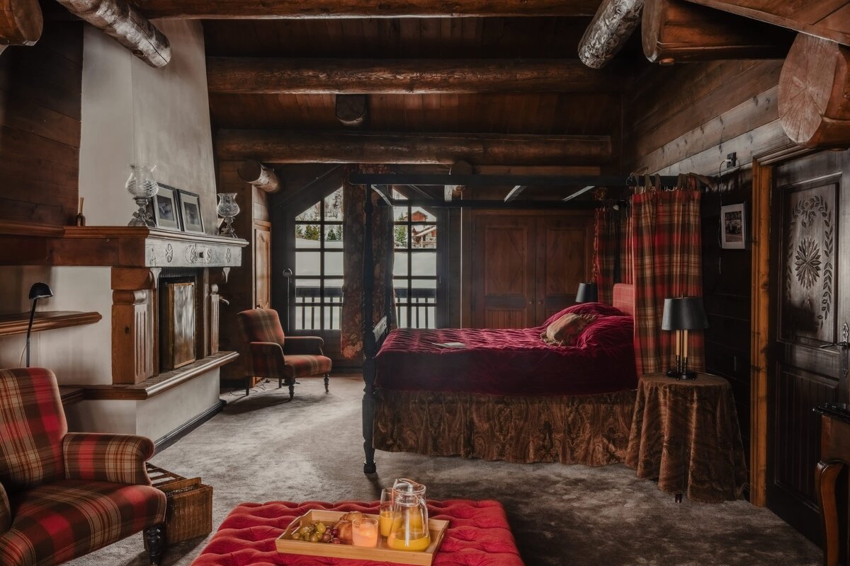 Cozy, rustic log cabin bedroom with a four-poster bed, fireplace, plaid chairs, and a snowy view from large windows.