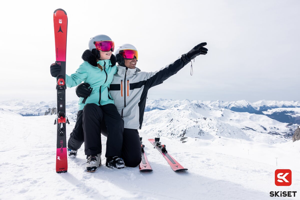 Two skiers posing on a snowy mountaintop, with one pointing towards the scenic view.