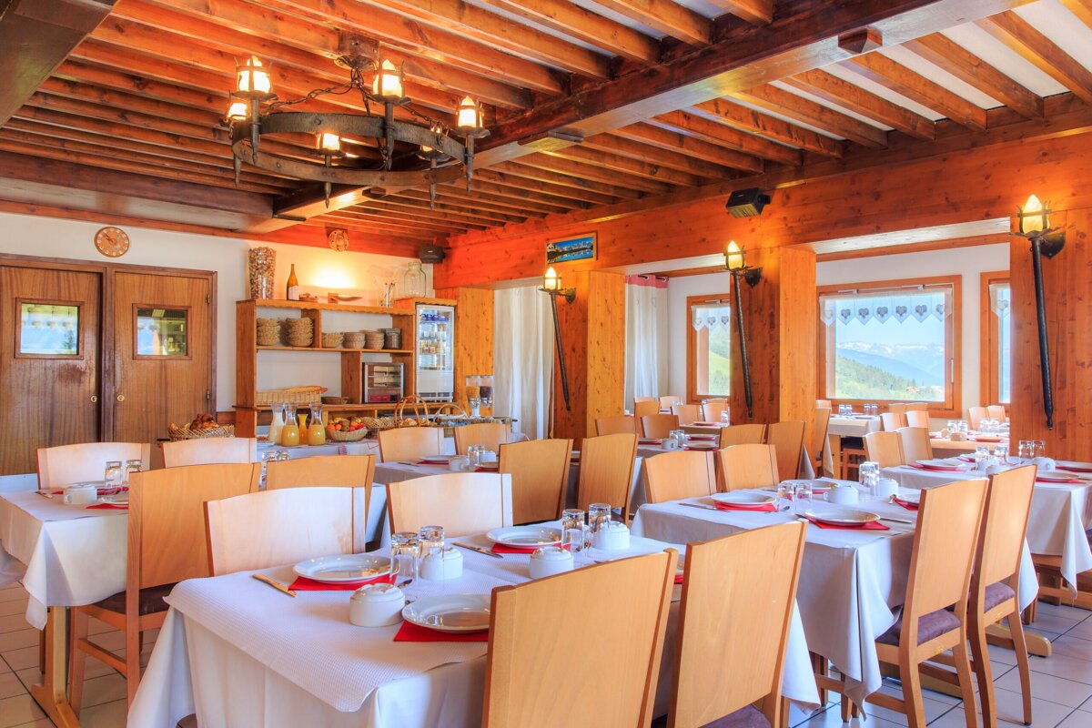 A rustic dining room with wooden ceilings, set tables, and a breakfast buffet. Large windows overlook a scenic mountain landscape, providing a warm, inviting atmosphere.
