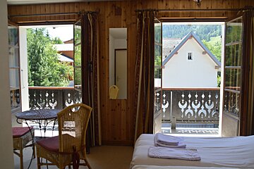Bedroom with twin balconies opening to a view of a white house, trees, and distant mountains. Wood-paneled walls, a bed, and wicker furniture inside.