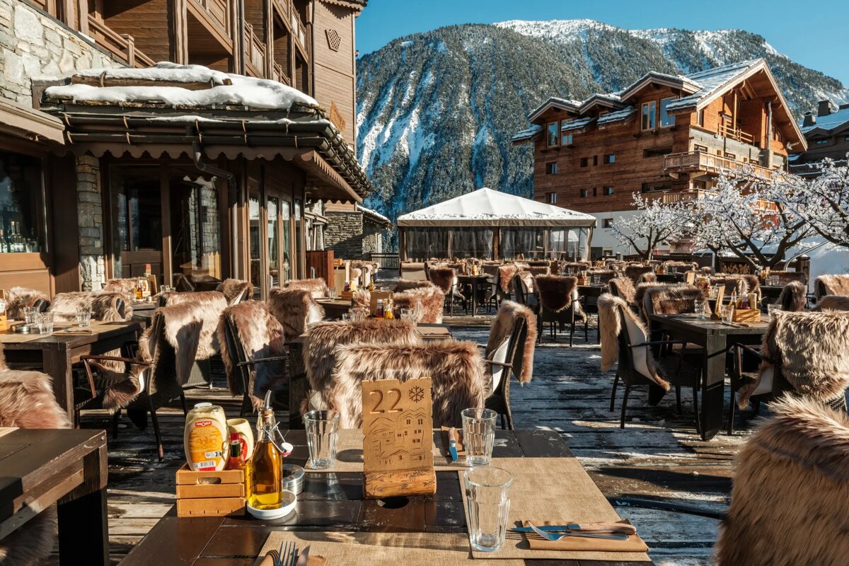A snowy outdoor restaurant patio with chairs draped in fur blankets, nestled among wooden buildings and a snow-capped mountain backdrop.