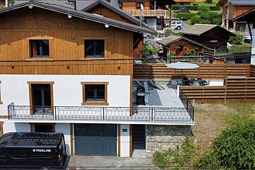 A multi-level chalet with a wood and white facade, featuring a balcony and a patio with outdoor furniture. A black van is parked, with other hillside homes.