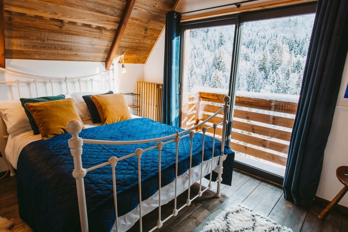 A cozy attic bedroom with a white bed, blue quilt, and golden pillows, featuring a large window overlooking a snowy forest.