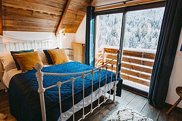 A cozy attic bedroom with a white bed, blue quilt, and golden pillows, featuring a large window overlooking a snowy forest.
