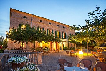 A charming stone building glows at dusk, its lighted windows overlooking an inviting outdoor patio with tables, chairs, and trees.