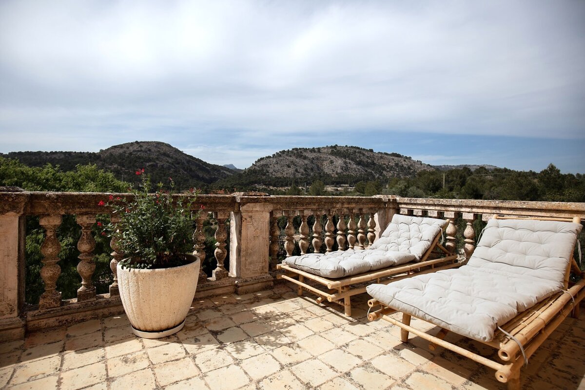 A sunny balcony with two bamboo loungers and a potted plant, offering a scenic view of rolling hills and distant mountains under a cloudy sky.