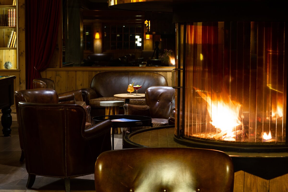 A cozy, dimly lit lounge features a roaring fireplace, plush brown leather seating, cocktails on a table, and bookshelves, creating a warm, inviting atmosphere.