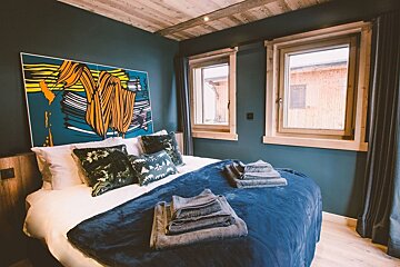 A cozy modern bedroom featuring dark blue walls, a wooden ceiling, a large bed with a blue throw, pop art, and twin windows.