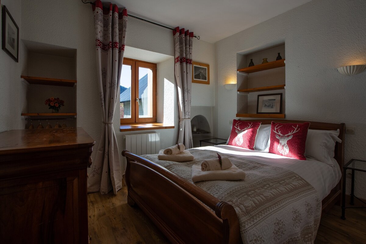 A cozy, rustic bedroom featuring a wooden bed with red deer-themed pillows, sunlit window, wooden shelves, and traditional furniture creating a warm atmosphere.