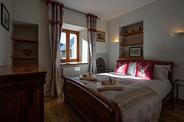 A cozy, rustic bedroom featuring a wooden bed with red deer-themed pillows, sunlit window, wooden shelves, and traditional furniture creating a warm atmosphere.