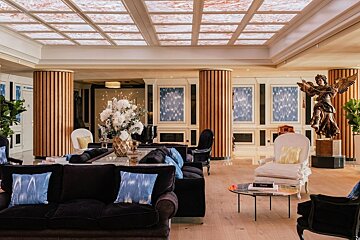 Luxurious lounge with dark sofas, white chairs, wooden columns, an ornate tiled ceiling, a grand floral arrangement, and an angel statue.