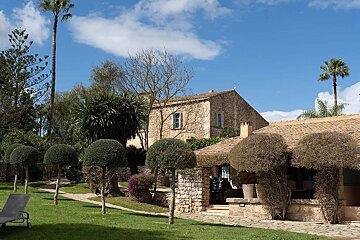 A stone house with a thatched roof is surrounded by trees and bushes