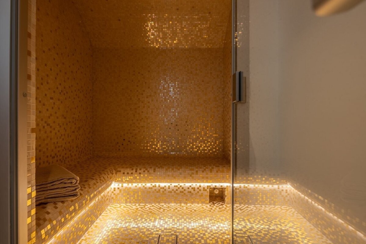 Luxurious steam room with shimmering gold mosaic tiles, a built-in bench with a towel, and warm LED accent lighting.
