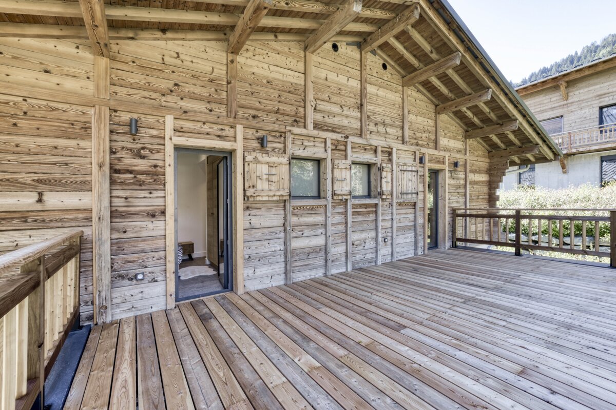 A spacious outdoor wooden deck extends from a rustic timber chalet, featuring doors, shuttered windows, and a railing against a hillside backdrop.
