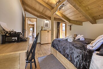 A cozy, rustic bedroom featuring a plush bed with grey bedding, exposed wooden beams, and a vintage typewriter on a desk. Natural wood tones dominate.