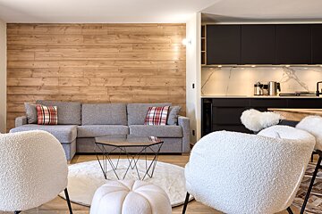 A cozy, modern open-plan living room and kitchen. Features a wood accent wall, gray sectional sofa, white chairs, and a sleek black kitchen with a marble backsplash.