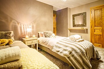 Cozy bedroom with two beds, soft lamplight, and plush faux fur throws in neutral tones. Features a rustic mirror, wooden door, and warm ambient glow.