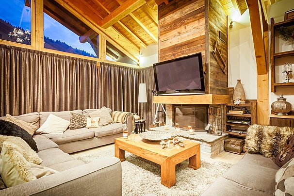 A warm, wood-paneled chalet living room with a comfy sectional sofa, fireplace, TV, and large windows offering a night mountain view.