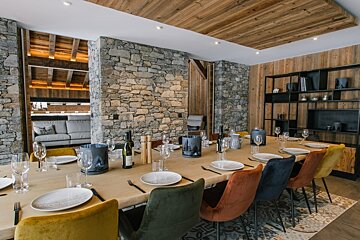 A chic, rustic dining room with a long table set with colorful chairs, stone walls, wooden ceilings, and a glimpse into a modern living space.