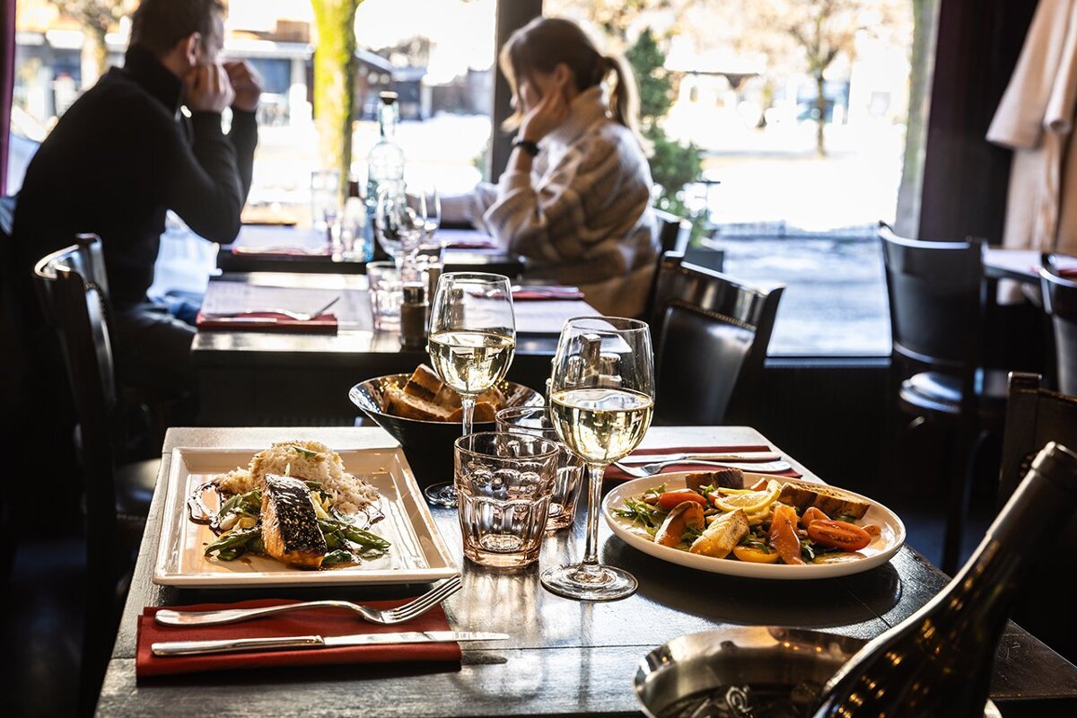 A couple sit at a table in a restaurant with plates of food and glasses of wine