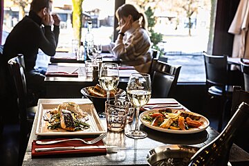A couple sit at a table in a restaurant with plates of food and glasses of wine