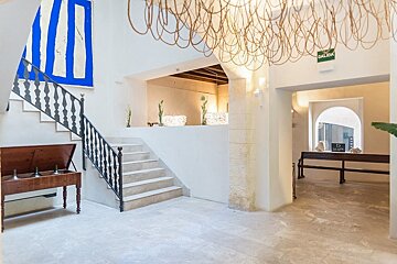 Elegant white lobby with a grand staircase, abstract blue art, and an intricate chandelier. A wooden bench and archway are also present.