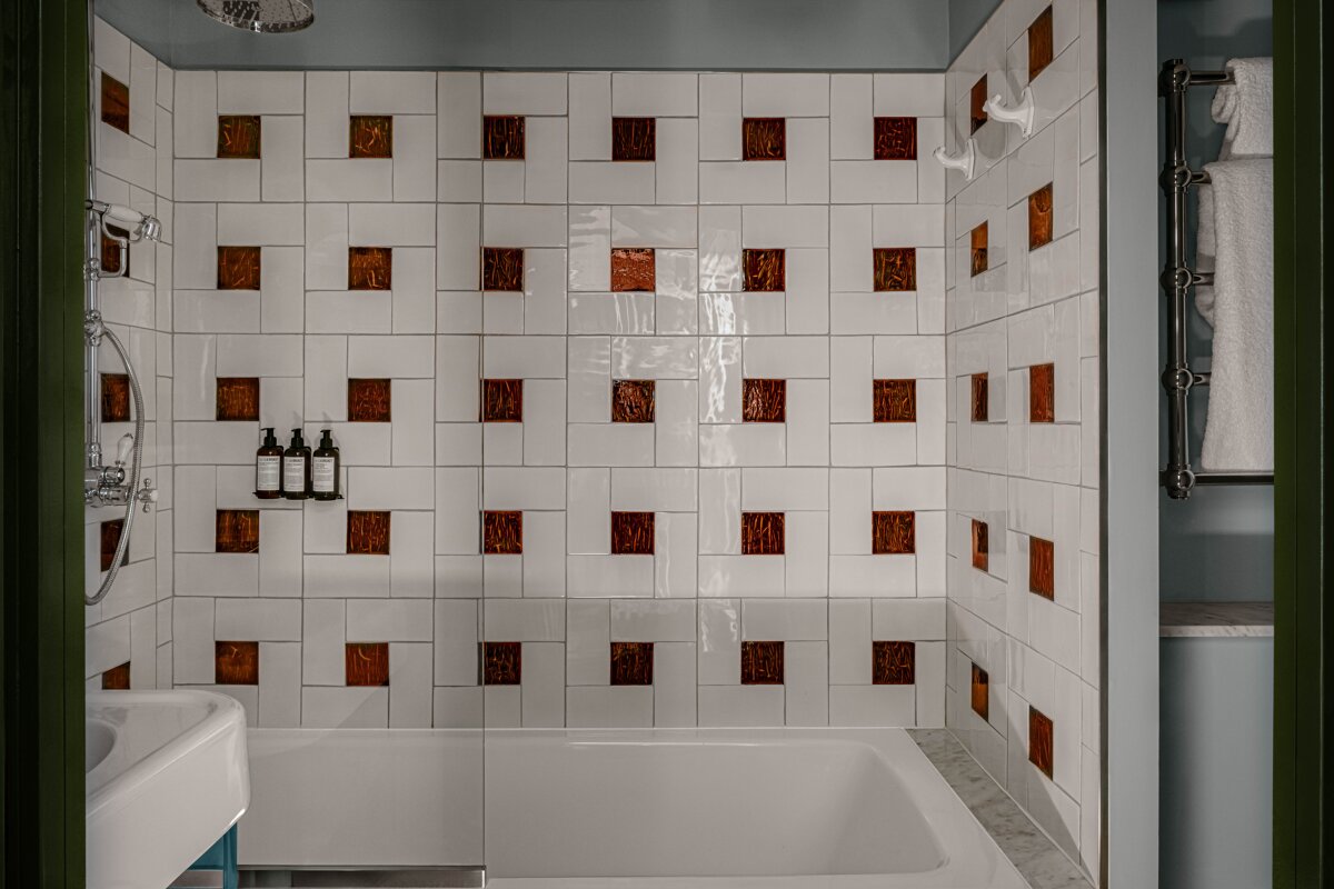 A modern bathroom features a white bathtub and shower. Walls are tiled white with square, textured copper-brown inserts, creating a unique pattern.