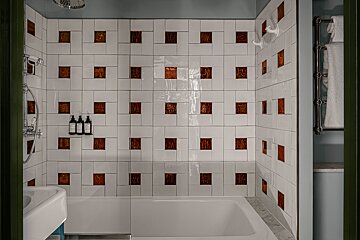 A modern bathroom features a white bathtub and shower. Walls are tiled white with square, textured copper-brown inserts, creating a unique pattern.