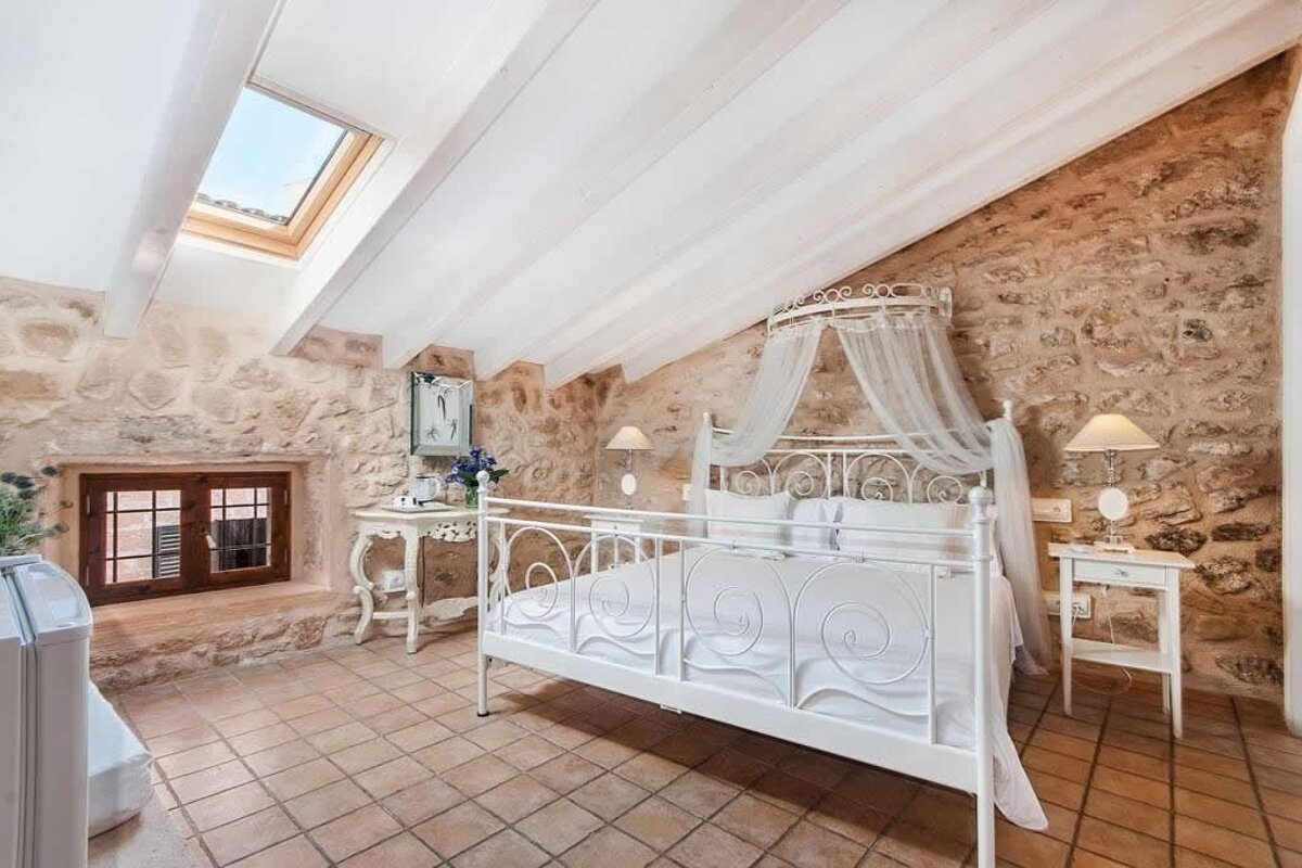 A rustic bedroom with stone walls, a white wrought-iron canopy bed with sheer drapes, terracotta tiled floor, and a skylight in the sloped ceiling.