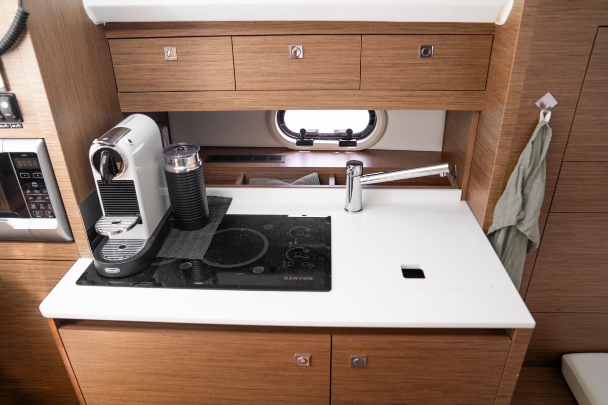 A modern boat galley features a Nespresso machine, electric stove, sink, microwave, and wooden cabinets, with a porthole window.