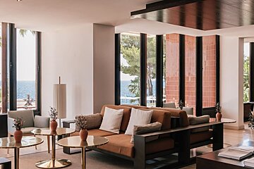 Modern lounge with burnt orange sofas, shiny tables, and large windows revealing an expansive sea view and lush trees.