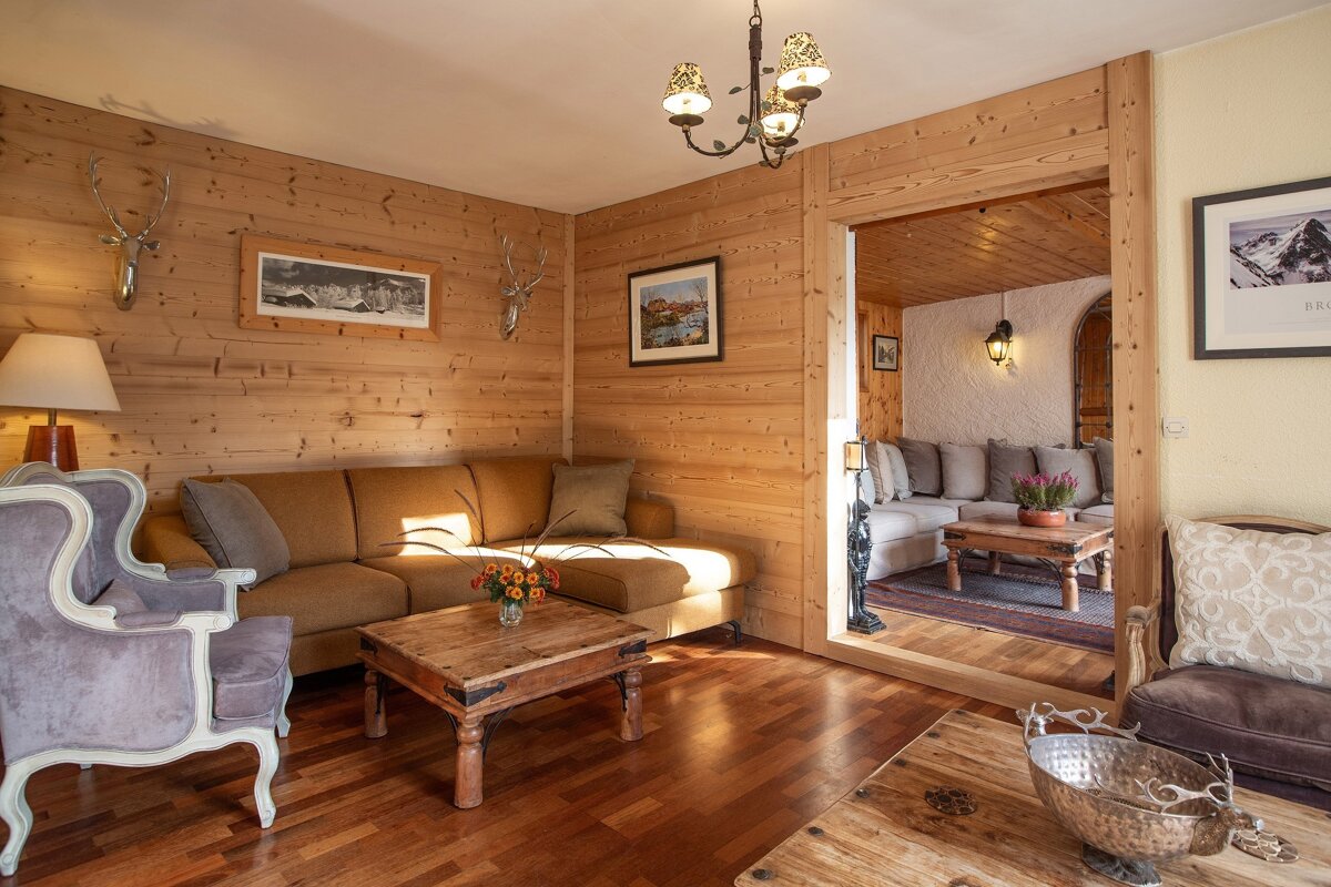 A cozy, wood-paneled living room with a brown sectional, rustic coffee tables, antler decor, and a chandelier, leading into another warm space.