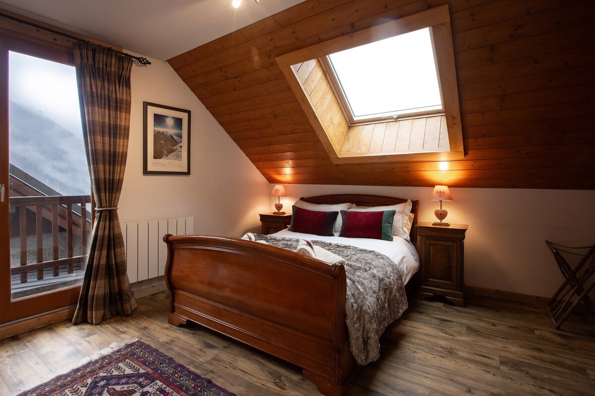 A cozy bedroom with a wooden sleigh bed, slanted wood ceiling, skylight, and a balcony door offering a mountain view. Warm lamps illuminate the space.