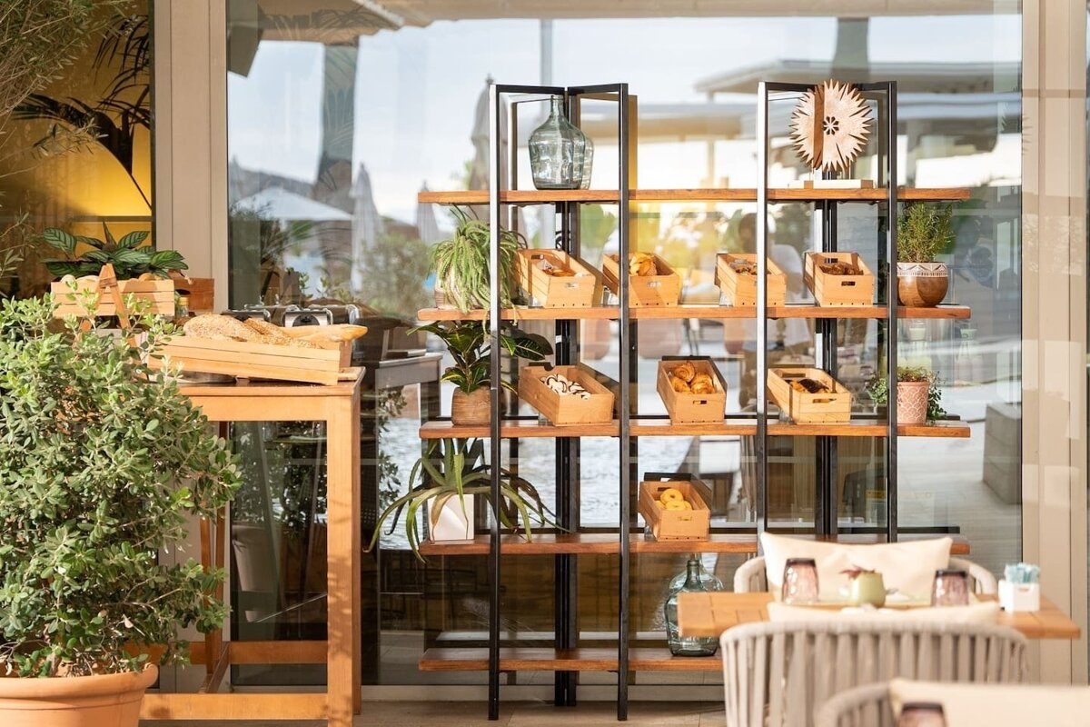 A multi-tiered shelf displays fresh pastries, bread, and potted plants in an inviting, light-filled space with glass windows reflecting the outdoors.