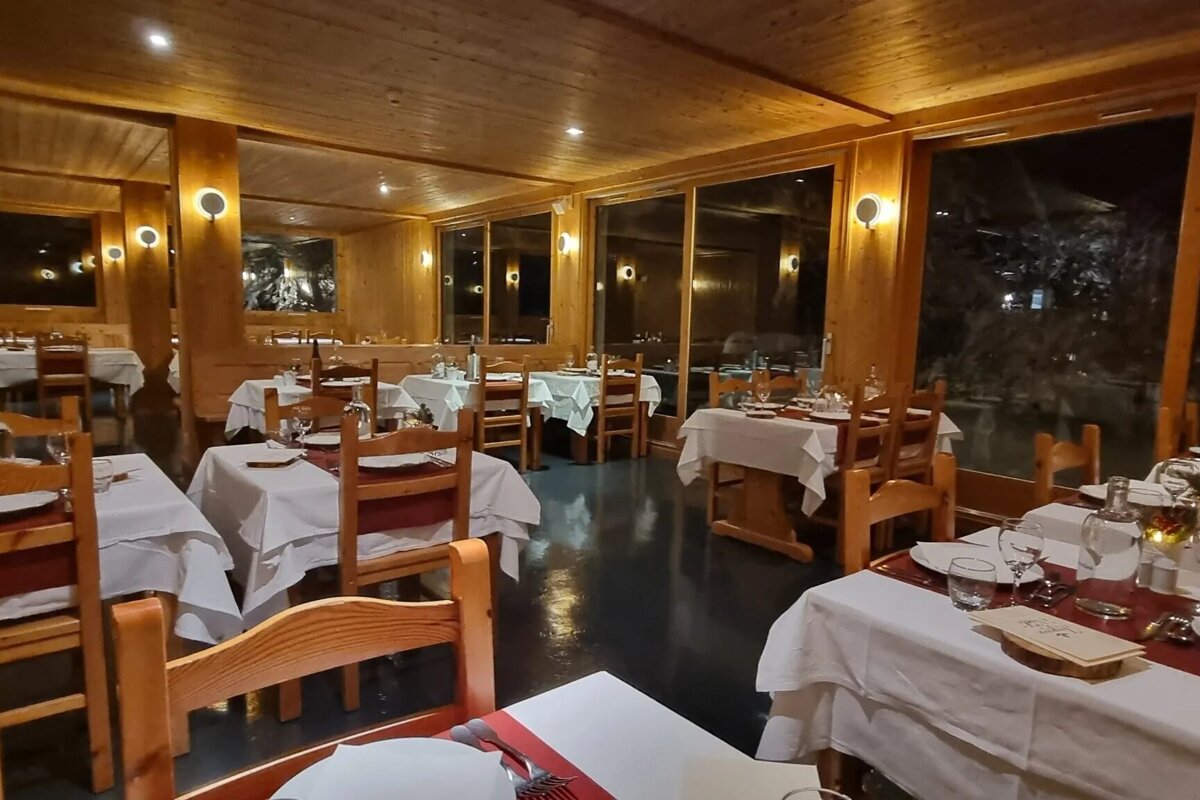A warmly lit, rustic wooden restaurant at night, featuring multiple tables set with white cloths and large windows looking out into the dark.