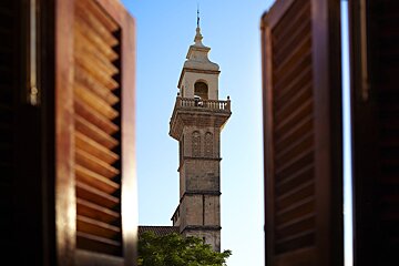 A clock tower is seen through a window with shutters open