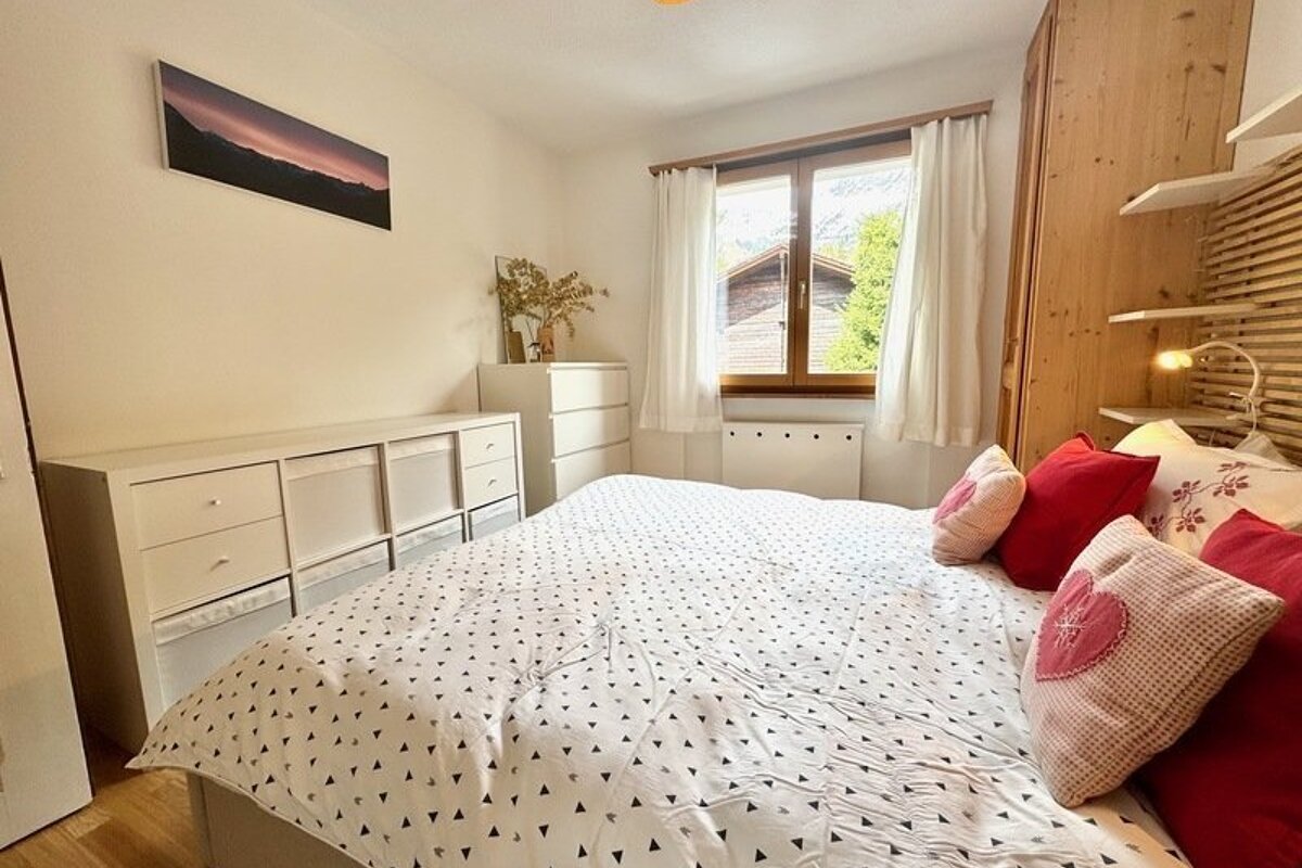 Bright bedroom featuring a patterned bed with red pillows, white dressers, and a window view of trees and a chalet.