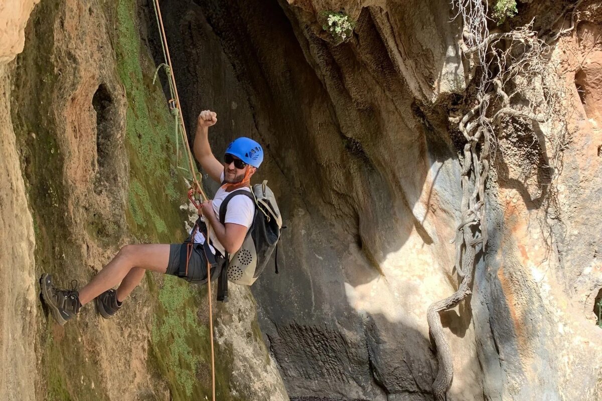A man wearing a blue helmet and sunglasses is climbing a rock wall