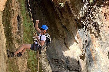 A man wearing a blue helmet and sunglasses is climbing a rock wall