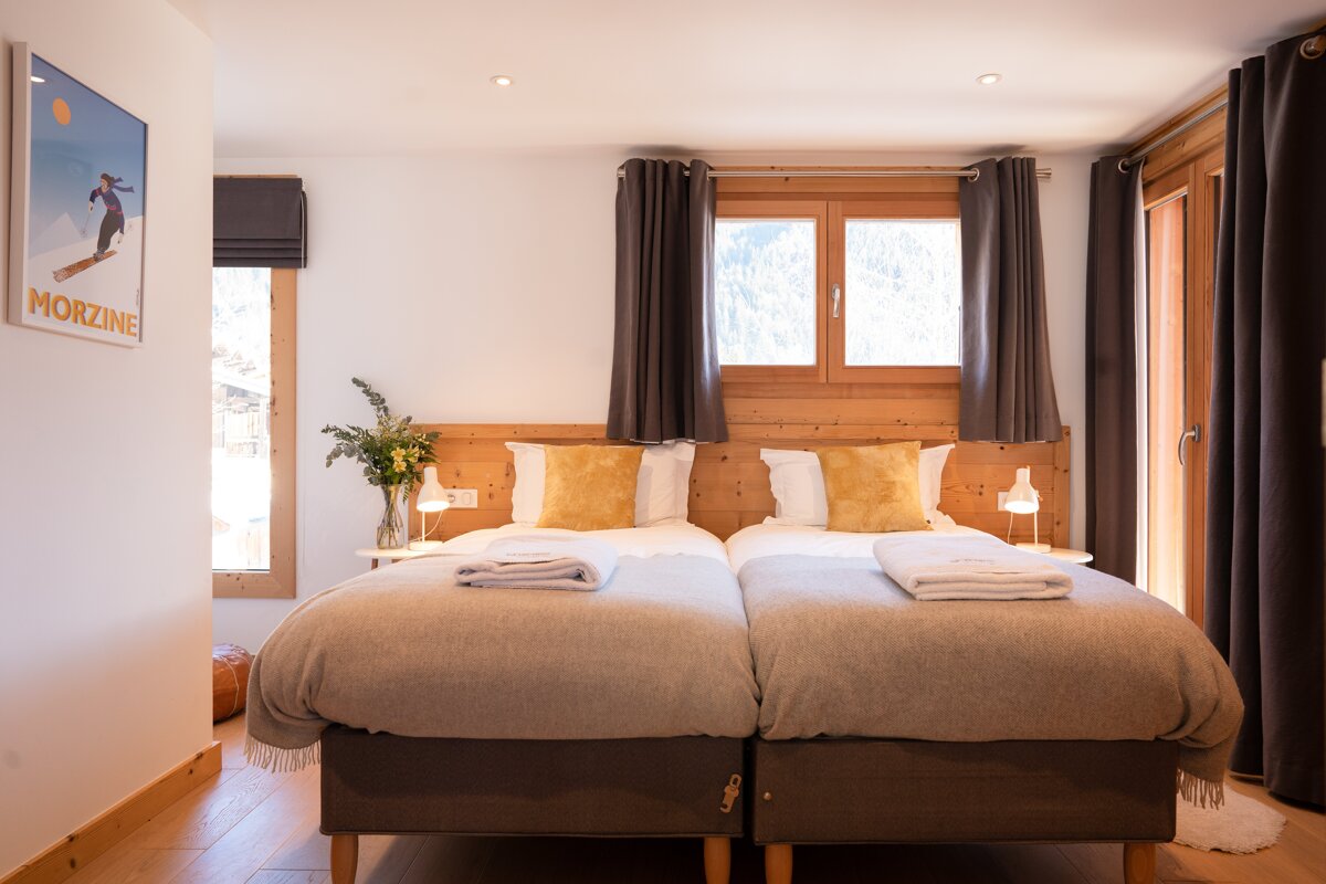 A bright, cozy bedroom with twin beds, wooden details, yellow pillows, and a mountain view. A ski poster adorns the white wall.