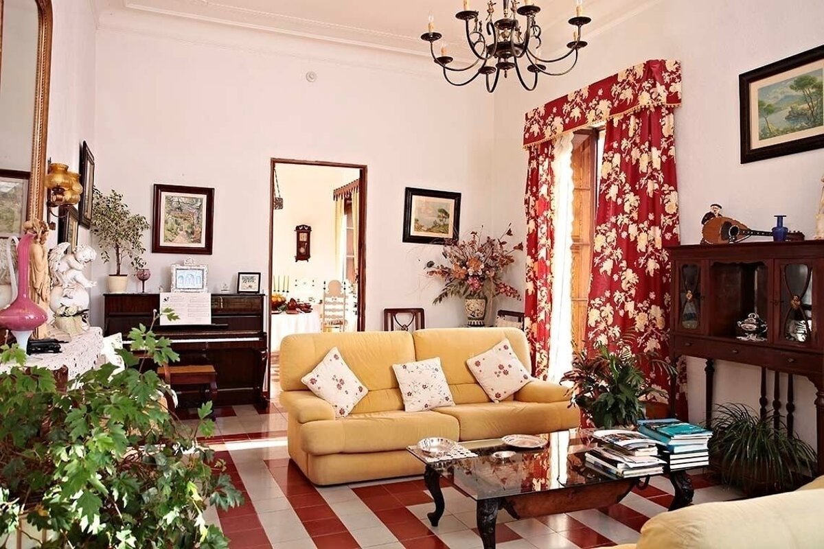 A cozy, traditionally decorated living room featuring a yellow sofa, red & white striped floor, red floral curtains, a piano, plants, and an ornate chandelier.
