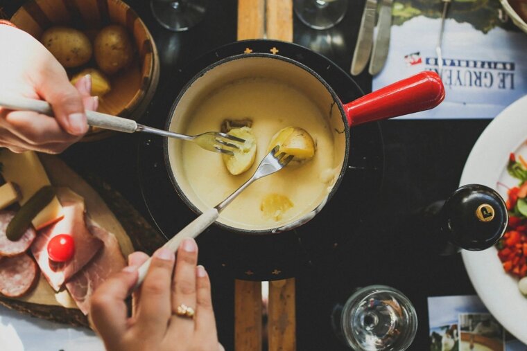 Two people dip potatoes into a creamy cheese fondue pot with forks. Meats, cheese, and salad are also on the dark table, creating a cozy shared meal.