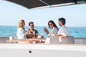 Four friends smiling and enjoying drinks around a table on a yacht, with the calm blue ocean in the background.